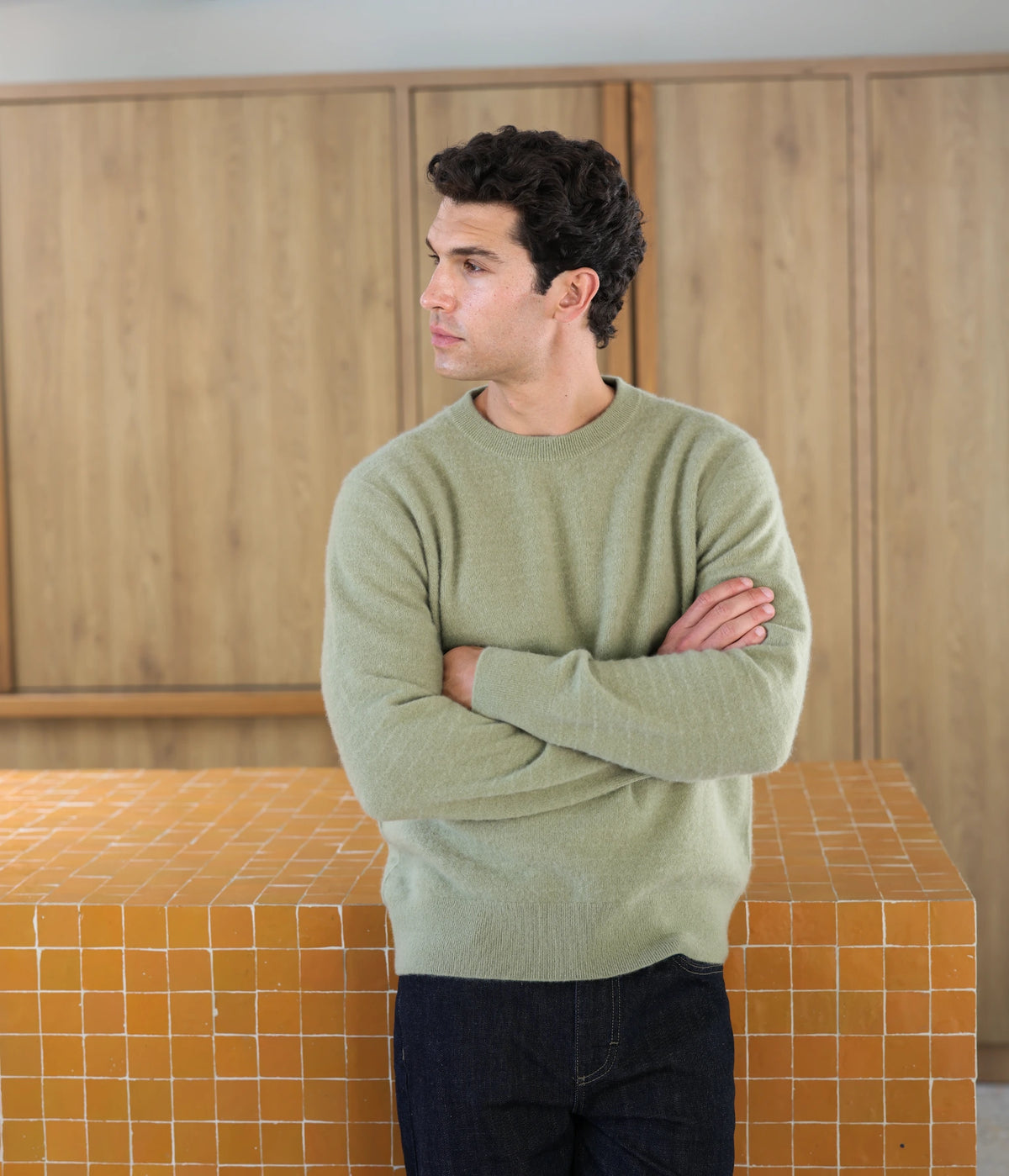 A man wearing a light sage green Uncle Purl crewneck sweater and dark jeans, standing with arms crossed in front of a wood-paneled wall and an orange tiled counter.