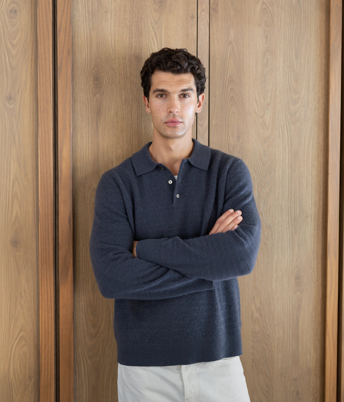 Uncle Purl Campaign. A man wearing a navy blue long-sleeve polo sweater and off-white trousers, standing with arms crossed in front of a vertical wood-paneled wall.
