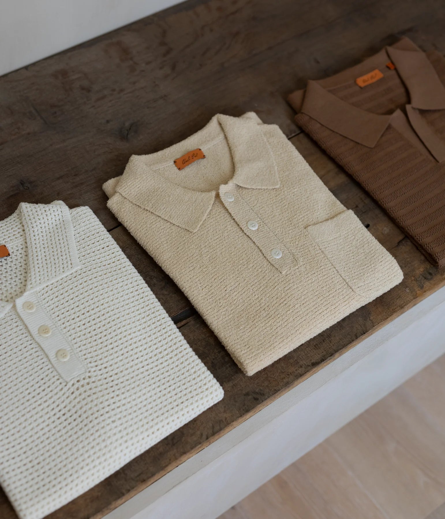 Three folded knit polo shirts arranged on a dark wooden surface: one cream, one warm beige, and one brown, each showing a small orange neck label.