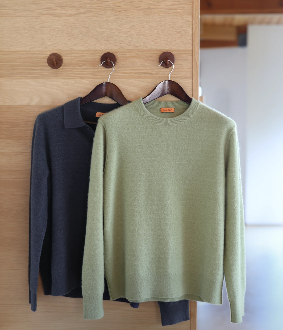 A light sage green crewneck sweater hangs on a wooden hanger in front of a dark charcoal gray polo sweater, both displayed on wooden pegs against a light wood-paneled wall.