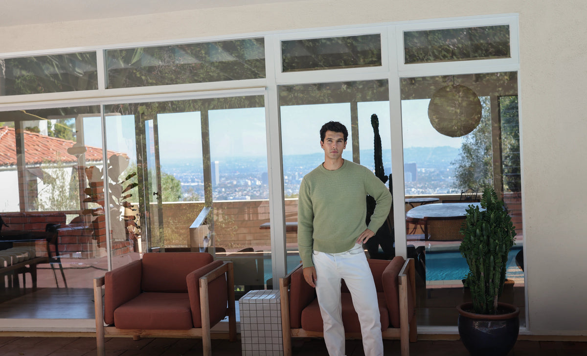 Uncle Purl Holiday Campaign. A man wearing a sage green long-sleeve crewneck sweater and white pants stands in a modern living room with large windows overlooking a city skyline and pool area.