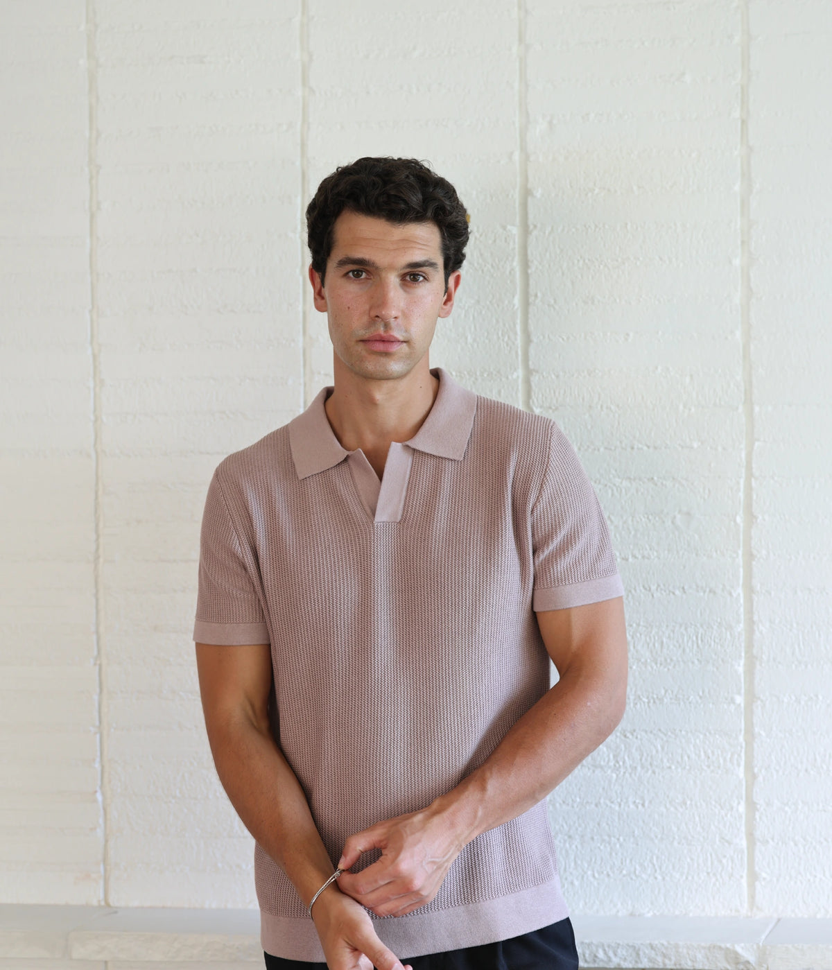 A man wearing a short-sleeve ribbed knit polo shirt in a dusty mauve-pink color, standing against a white textured wall.