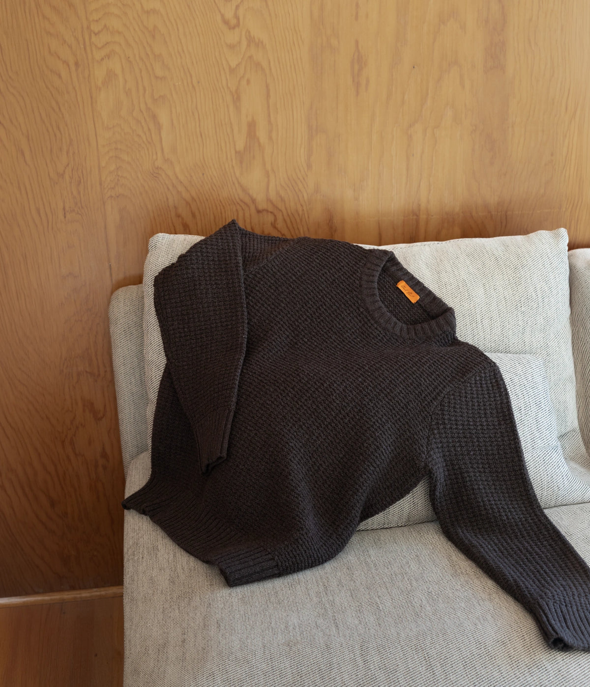 Uncle Purl Merino Waffle Sweater in brown. A dark brown textured knit sweater with a small orange tag, draped over a light beige upholstered armchair in front of a wooden wall.